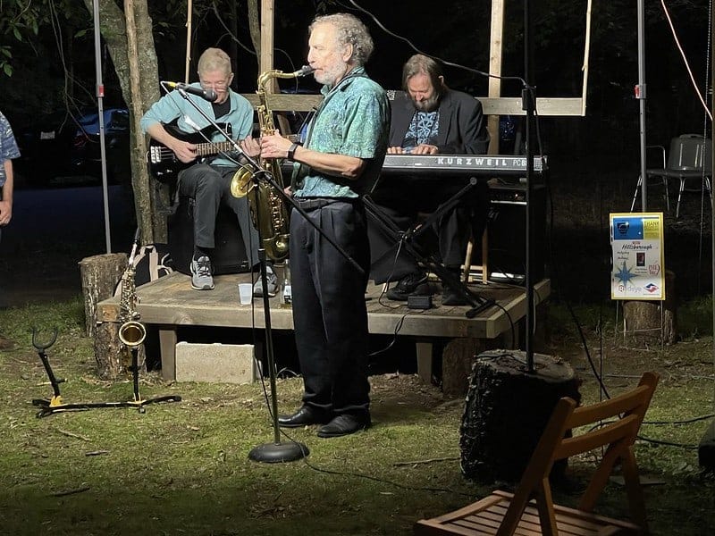 David Kreimer singing into a microphone outdoors at night with an alto sax on a strap, Rick Keena on bass guitar behind him and Marc Puricelli at a Kurzweil keyboard.